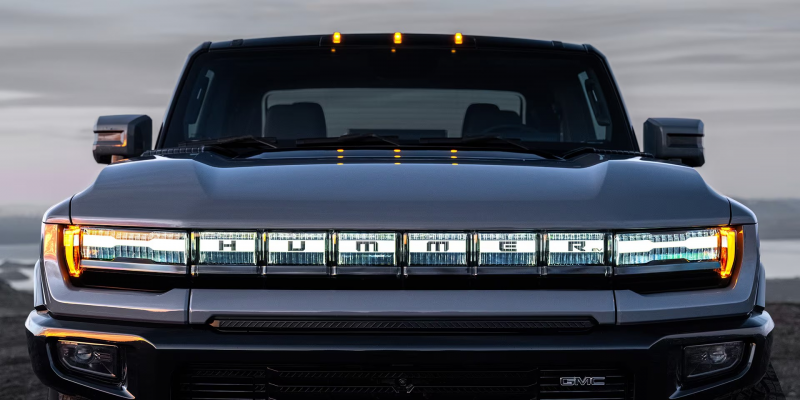 Front view of the 2025 GMC Hummer EV, highlighting its illuminated Hummer grille and rugged design with amber marker lights glowing against a twilight backdrop