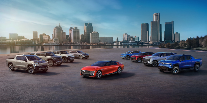 Lineup of 2025 General Motors electric vehicles, including trucks and sedans, showcased on a paved surface with a city skyline and waterfront in the background under a clear blue sky.